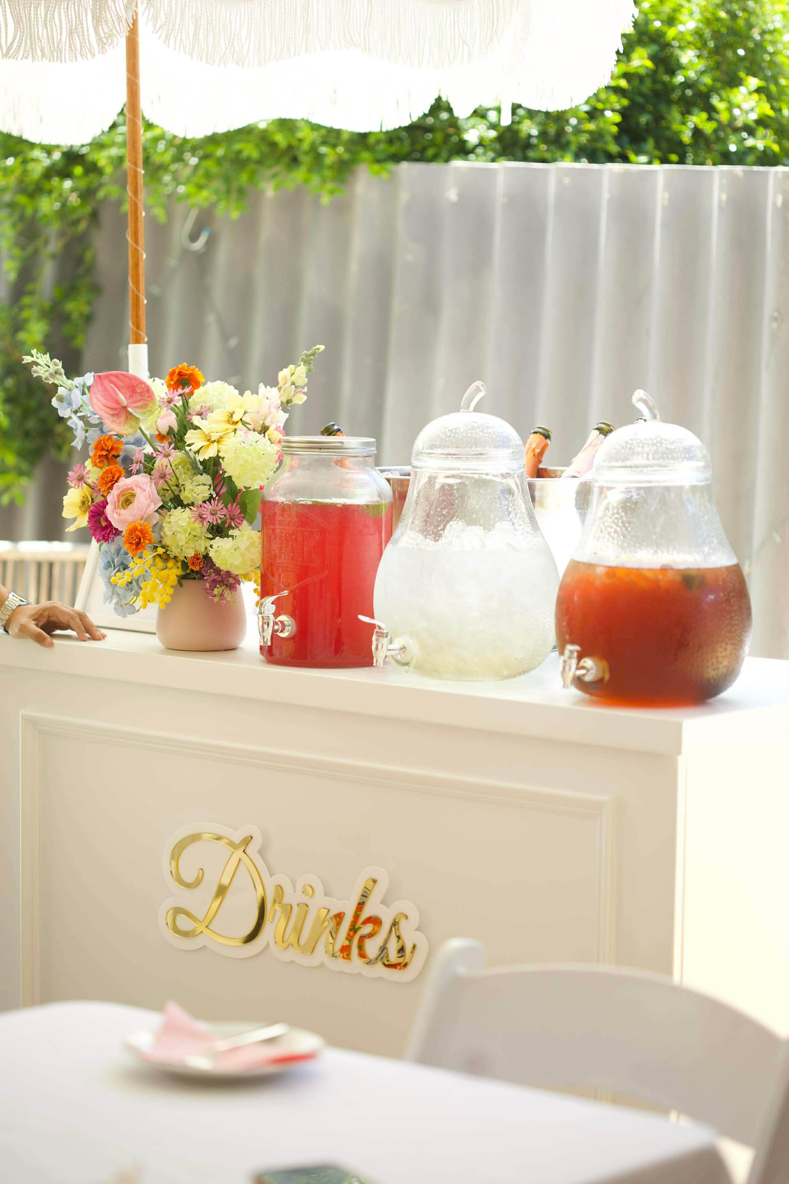 White drinks cart with glass dispensers, colourful flowers, and gold Drinks lettering